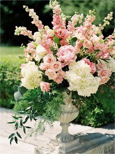 a vase filled with pink and white flowers sitting on top of a wooden table next to bushes