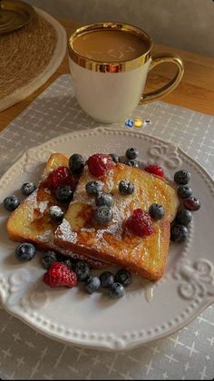 two pieces of french toast on a plate with berries and powdered sugar next to a cup of coffee