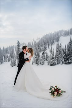 a bride and groom kissing in the snow with pine trees behind them on their wedding day