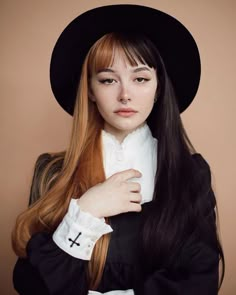 a woman with long hair wearing a black hat and white collared shirt, standing in front of a brown background