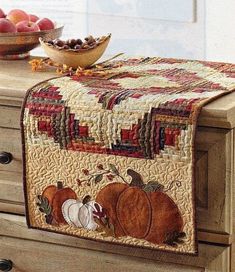 a quilted table runner with pumpkins and apples in the background on a dresser