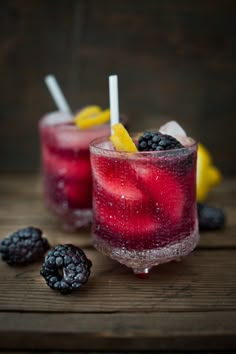 two glasses filled with fruit and ice sitting on top of a wooden table next to blackberries