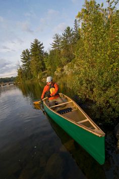 a man in an orange life jacket paddling a green canoe on the water near some trees