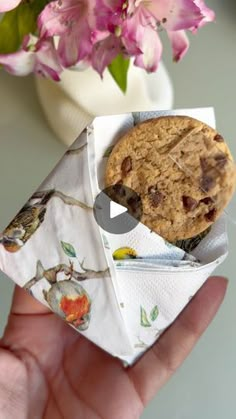 a hand holding an origami chocolate chip cookie and pink flowers in the background