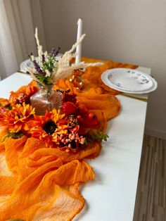 an arrangement of flowers and candles on a long table with orange fabric draped over it