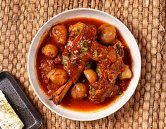 a white bowl filled with meat and potatoes on top of a table next to a piece of bread