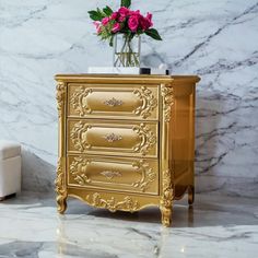 a gold chest of drawers with flowers in a vase on top and marble walls behind it