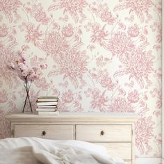 a white bed sitting under a pink wallpaper next to a wooden chest of drawers