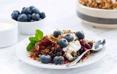 a white plate topped with fruit and granola next to a bowl of blueberries