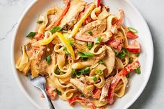 a white plate topped with pasta covered in chicken and peppers next to a fork on a marble surface