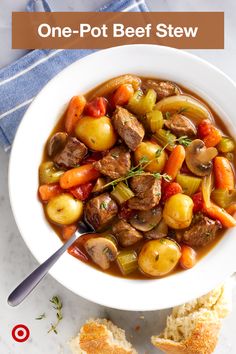 a white bowl filled with beef stew and potatoes on top of a table next to bread