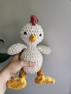 a hand holding a crocheted chicken stuffed animal in front of a gray wall