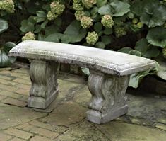 a stone bench sitting on top of a brick floor next to green plants and bushes