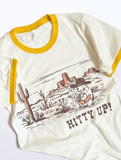 Let's go girls!  Our instant classic Kitty Up design is ready for summer! This super cute retro style tee is made with soft cotton and features our famous cowboy cat. A must have for desert dwellers and city citizens alike! Unisex sized and printed on a cream color tee with yellow accent binding on the sleeves and neckline.  Available in sizes XS S M L XL Copyright 2024 The Coin Laundry Fun Graphic Tees, Coin Laundry, Tshirt Design Inspiration, Willow Creek, Shirt Design Inspiration, Funny Tee Shirts, Cowgirl Western, Ringer Tee, Cow Girl