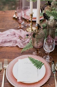 the table is set with pink plates, silverware and greenery in vases