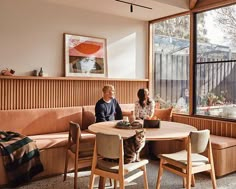 two people sitting at a round table in front of large windows with wood paneling