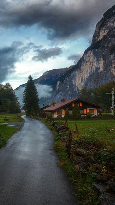 an empty road in the middle of a grassy area with mountains in the background and houses on either side