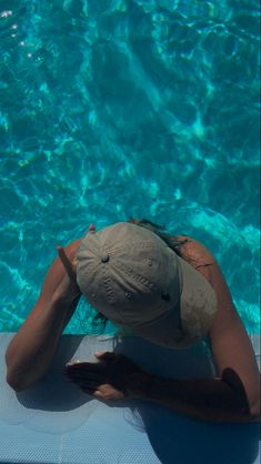 a woman in a hat sitting on the edge of a swimming pool with her hands behind her head