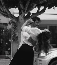 black and white photograph of two people dancing in front of a car on the street