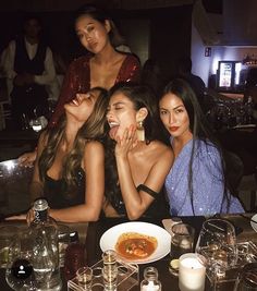 three women sitting at a table with food and drinks in front of them, posing for the camera