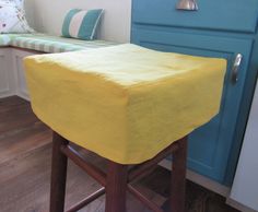 a stool with a yellow cover on it in front of a blue cabinet and window