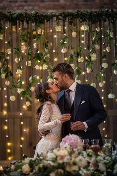 a bride and groom kissing in front of a table with flowers hanging from the ceiling