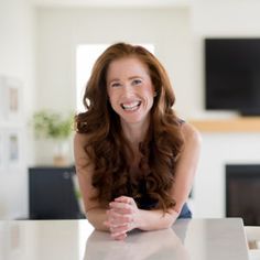 a woman sitting at a table with her arms crossed and smiling for the camera, in front of a fireplace