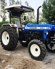 a blue and white tractor parked on top of a dirt covered field with trees in the background