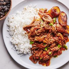 a white plate topped with meat, rice and beans next to a bowl of beans