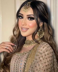 a woman with long brown hair wearing a gold dress and jewelry on her head, standing in front of a white wall