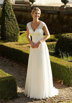 a woman in a white dress standing next to some hedges and bushes with her hands on her hips