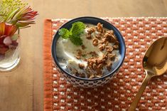 a bowl of cereal on top of a table next to a spoon and flower vase