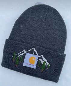 a grey beanie hat with a yellow c logo on the front and mountains in the background