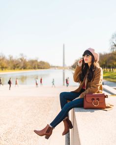 a woman sitting on top of a cement wall next to a lake and holding a brown purse