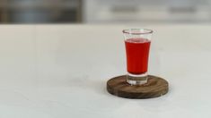 a glass filled with red liquid sitting on top of a white table next to a wooden coaster