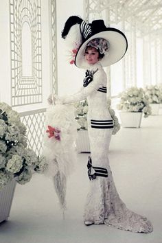 a woman in a white dress and black hat holding an umbrella with flowers behind her