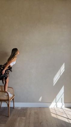 a woman sitting on top of a wooden chair in front of a wall with a window