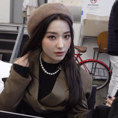 a woman sitting at a table with a laptop in front of her, wearing a hat and pearls necklace