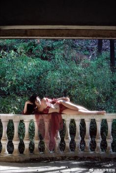 a woman laying on top of a white fence next to green bushes and trees in the background