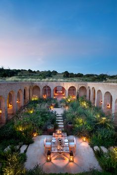 an outdoor dining area is lit up with candles and greenery at dusk, surrounded by stone arches