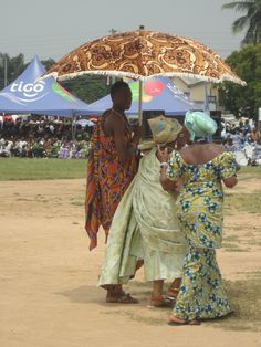 three women standing under an umbrella on a dirt field with people in the background watching