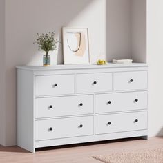 a white dresser in a room with a plant and pictures on the wall above it