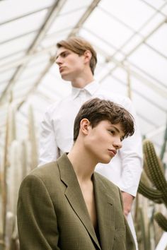 two men standing in a greenhouse with cacti and cactus behind them, one wearing a suit