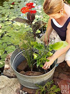 a woman kneeling down next to a metal bucket filled with water plants and other flowers