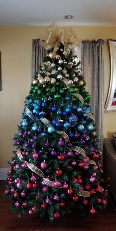 a brightly colored christmas tree in a living room decorated with ornaments and ribbon on the top