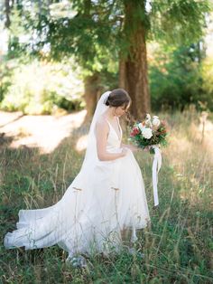 a woman in a wedding dress holding a bouquet and looking down at her hand while standing in the grass