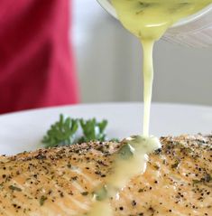 a person pouring sauce onto a piece of fish on a white plate with parsley