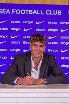 a man sitting at a desk in front of a blue sign with the words sea football club on it