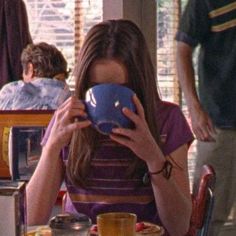 a woman sitting at a table holding a blue bowl in front of her face and covering her eyes