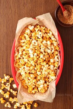 a red bowl filled with popcorn next to a small bowl of seasoning on top of a wooden table